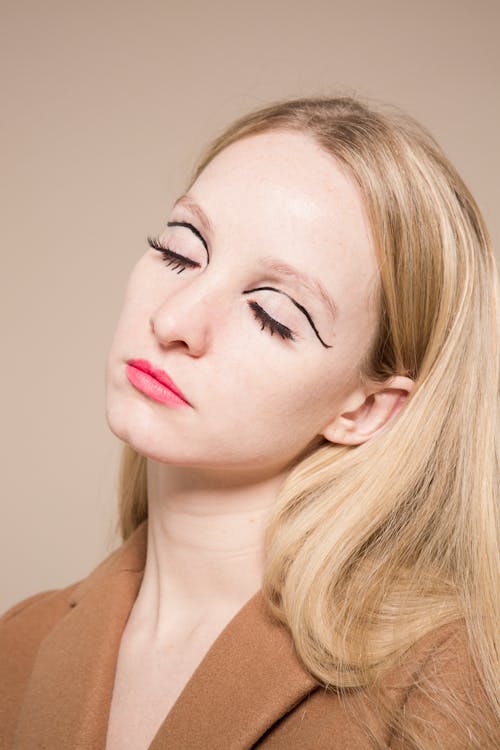 Tranquil stylish female with extraordinary makeup and eyes closed on beige background of studio