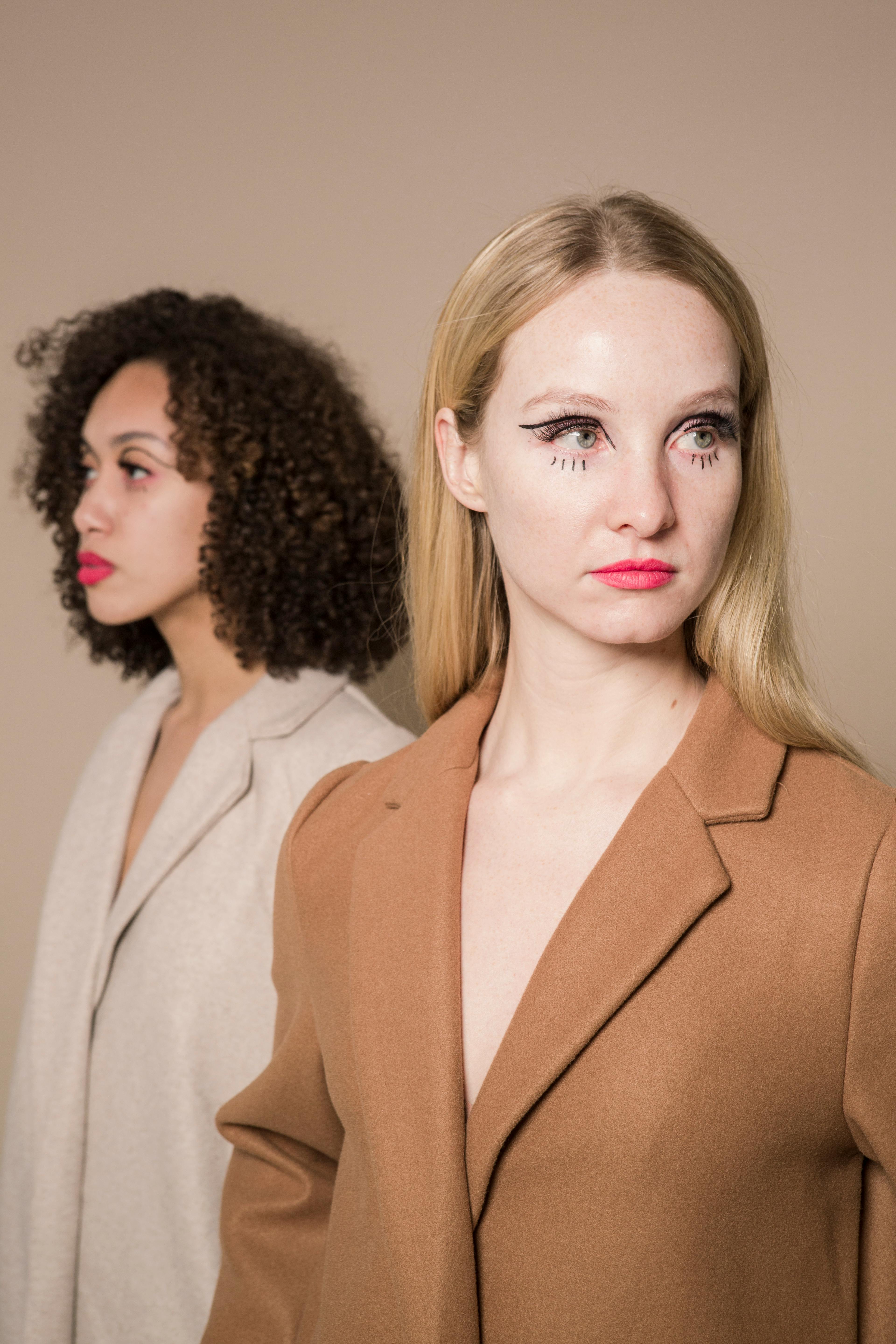 Slender multiethnic friends in trendy elegant coats quarreling and looking away on beige background of studio