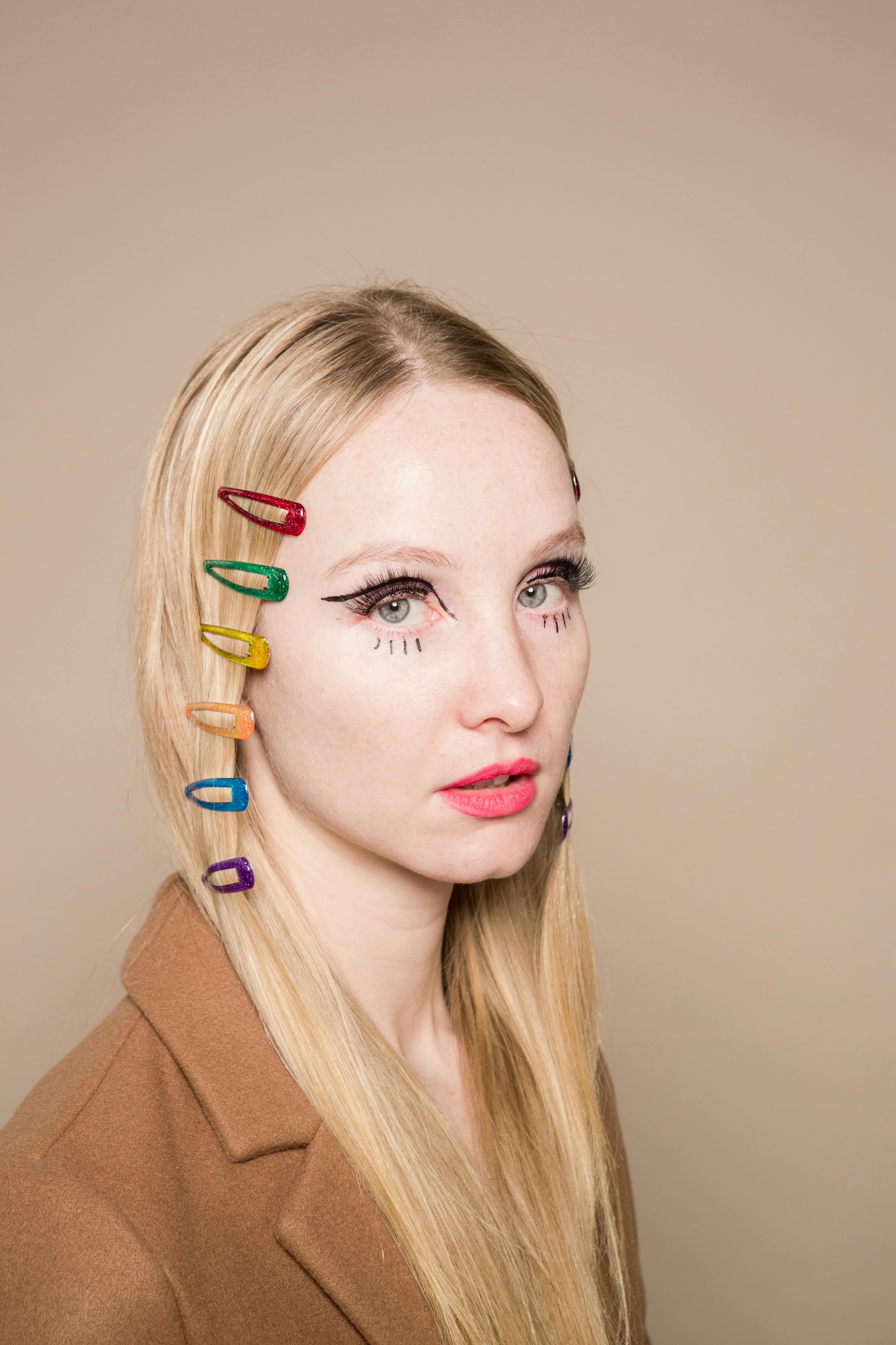 Pensive young woman with blond hair with multicolored hairpins and creative makeup looking at camera against beige background in studio