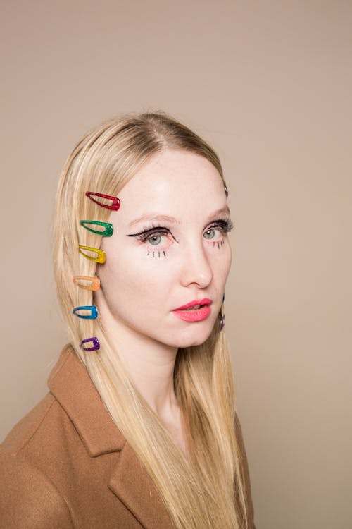 Pensive young woman with blond hair with multicolored hairpins and creative makeup looking at camera against beige background in studio