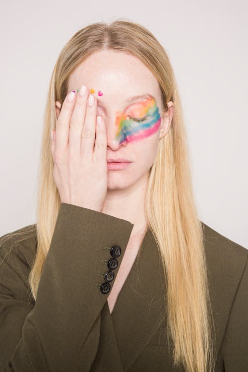 Calm female with long blond hair and colorful creative makeup in jacket standing with closed eyes and covering face against light background in studio