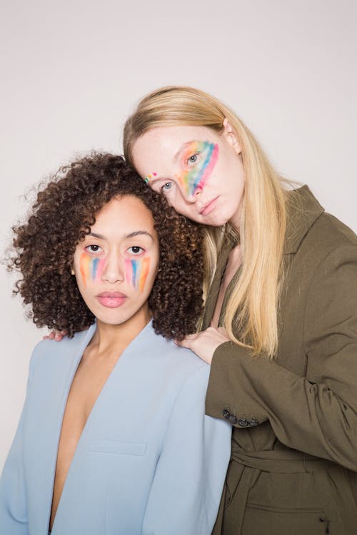 Serious multiracial women with colorful makeup as sign of LGBTQ in jackets looking at camera in studio with beige background