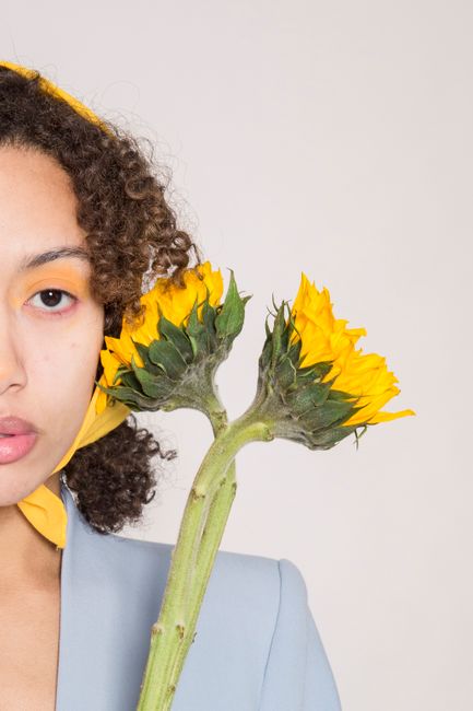 Confident woman with brown curly hair in yellow headscarf and stylish jacket holding blooming sunflower and looking at camera against light background