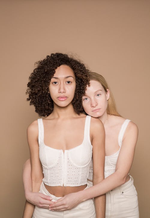 Diverse gentle women in white outfits hugging and looking at camera on beige background of studio