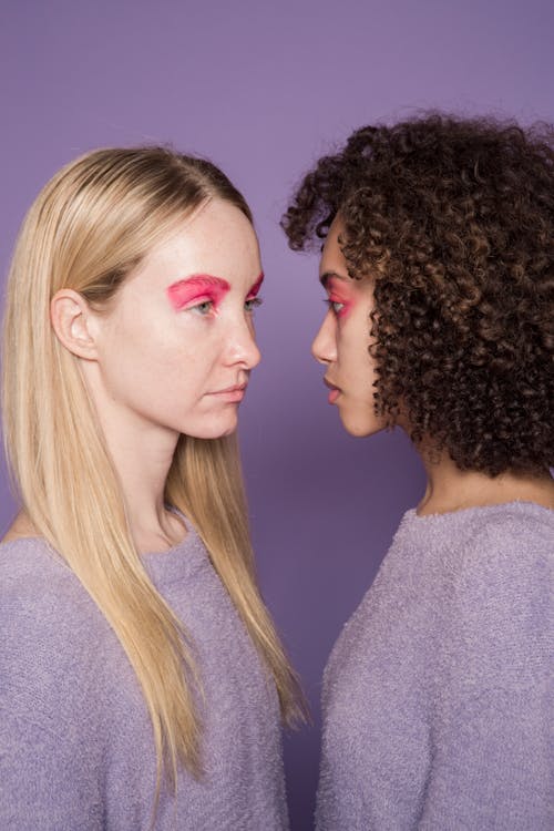 Conflicting multiethnic crop friends in soft pullovers and eccentric makeup on purple background of studio