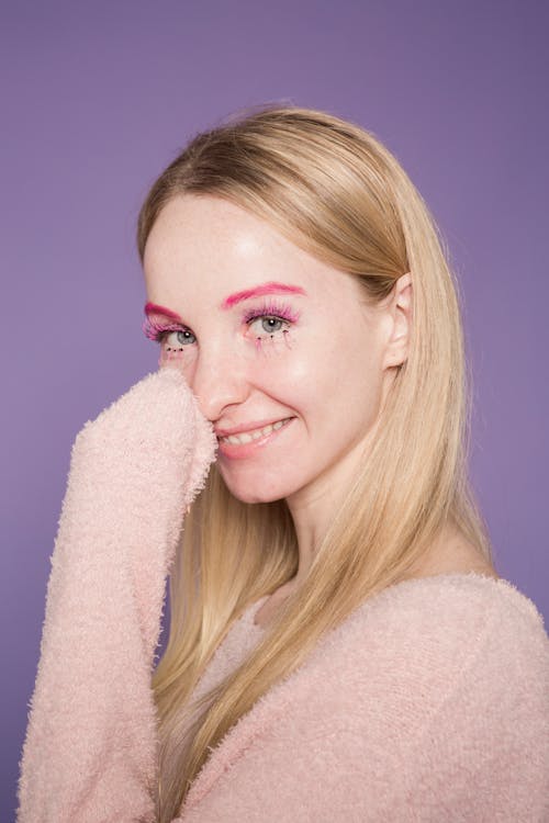 Glad female with bright pink eccentric makeup smiling and looking at camera on purple background