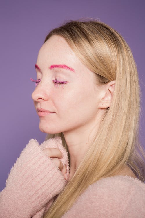 Gentle female with fair long hair and eyes closed in pink warm sweater on purple background