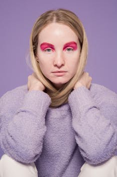 Portrait of a young woman with striking pink eye makeup, wearing a lavender sweater against a purple backdrop.