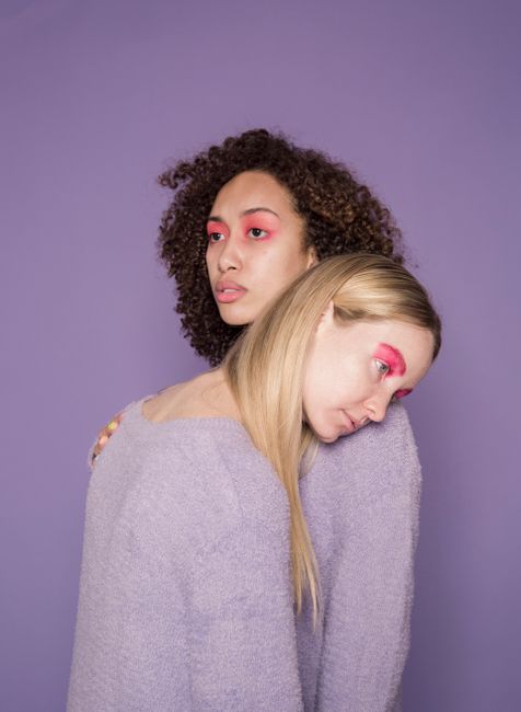 Serious young multiethnic female friends in sweaters with colorful pink makeup leaning on shoulder on violet background in studio