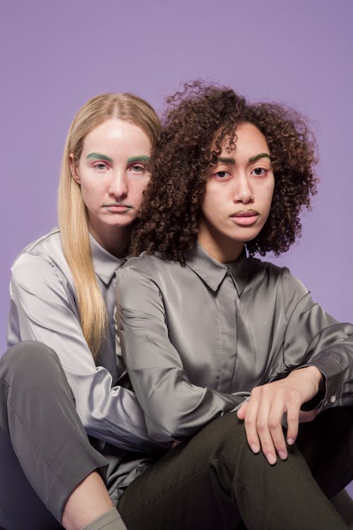 Serious young multiethnic female friends in shirts looking at camera while sitting and cuddling on violet background in studio