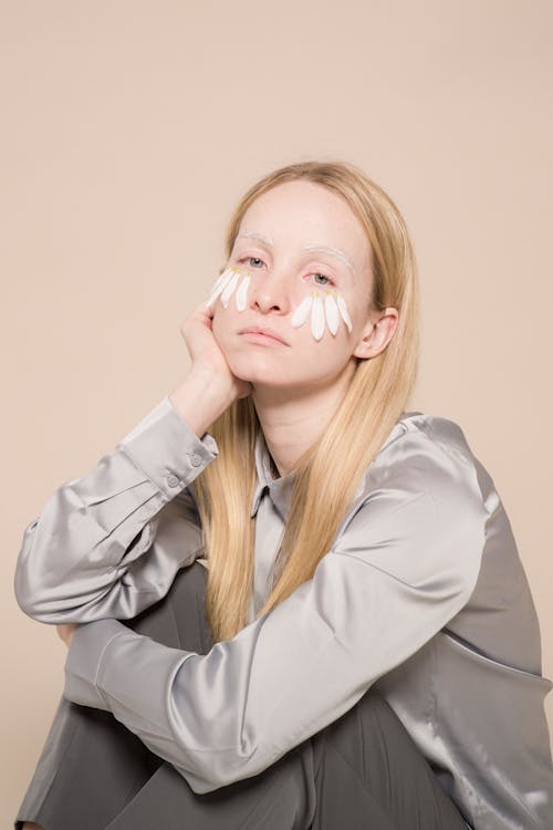 Tired young woman in shirt with white flower petals on face sitting with hand on face in bright studio while looking at camera on beige background