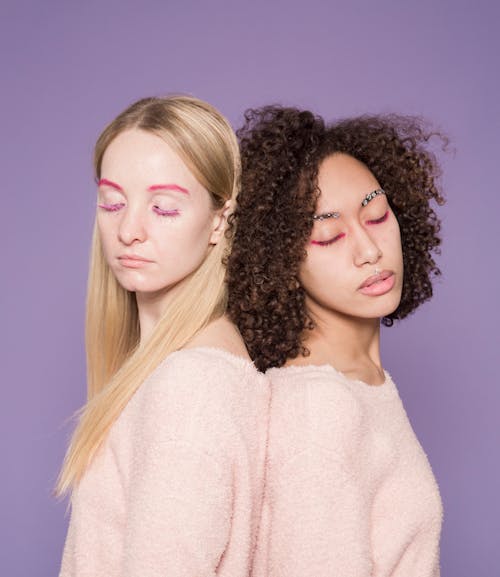 Multiracial tranquil friends in pink soft pullovers leaning backs on purple background of studio