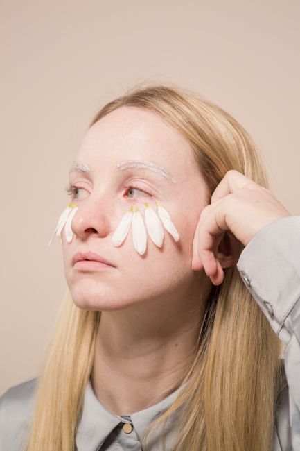 Young female with delicate white petals of chamomile on face looking away on beige background