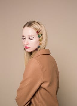 Emotionless young female in brown jacket with colorful makeup on face and barrettes in hair with closed eyes in bright studio on beige background