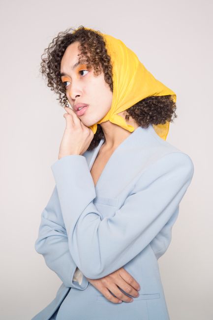 Serious young ethnic woman in blue jacket and yellow bandana on head with colorful makeup while looking away on gray background in light studio