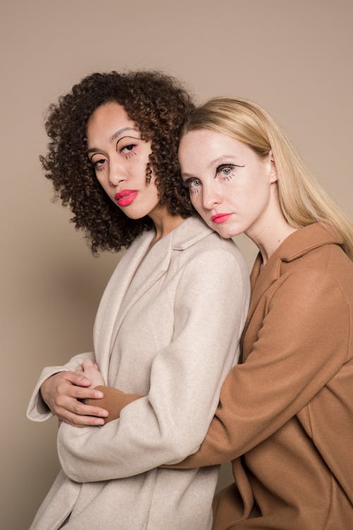 Confident multiracial female models with bright lipstick and colorful eyeliner hugging and looking at camera against beige background in studio
