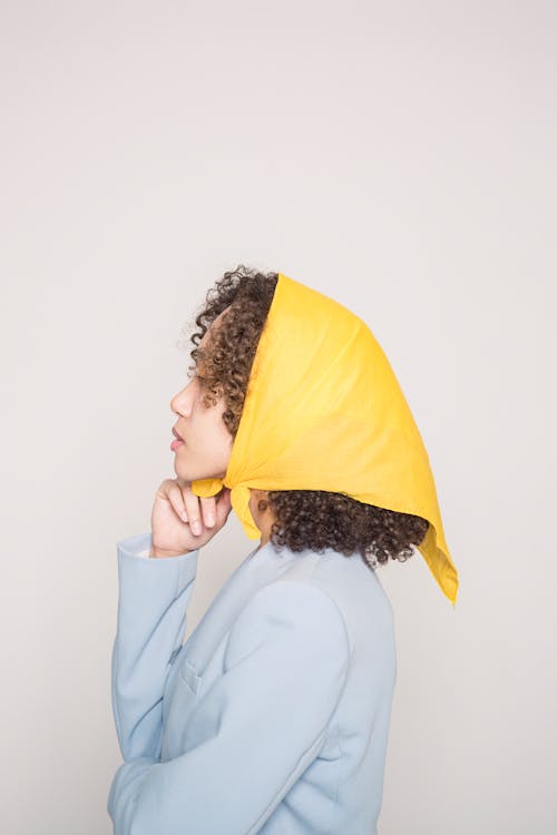 Side view of elegant ethnic female with black hair wearing trendy yellow headwear standing against gray background in modern studio