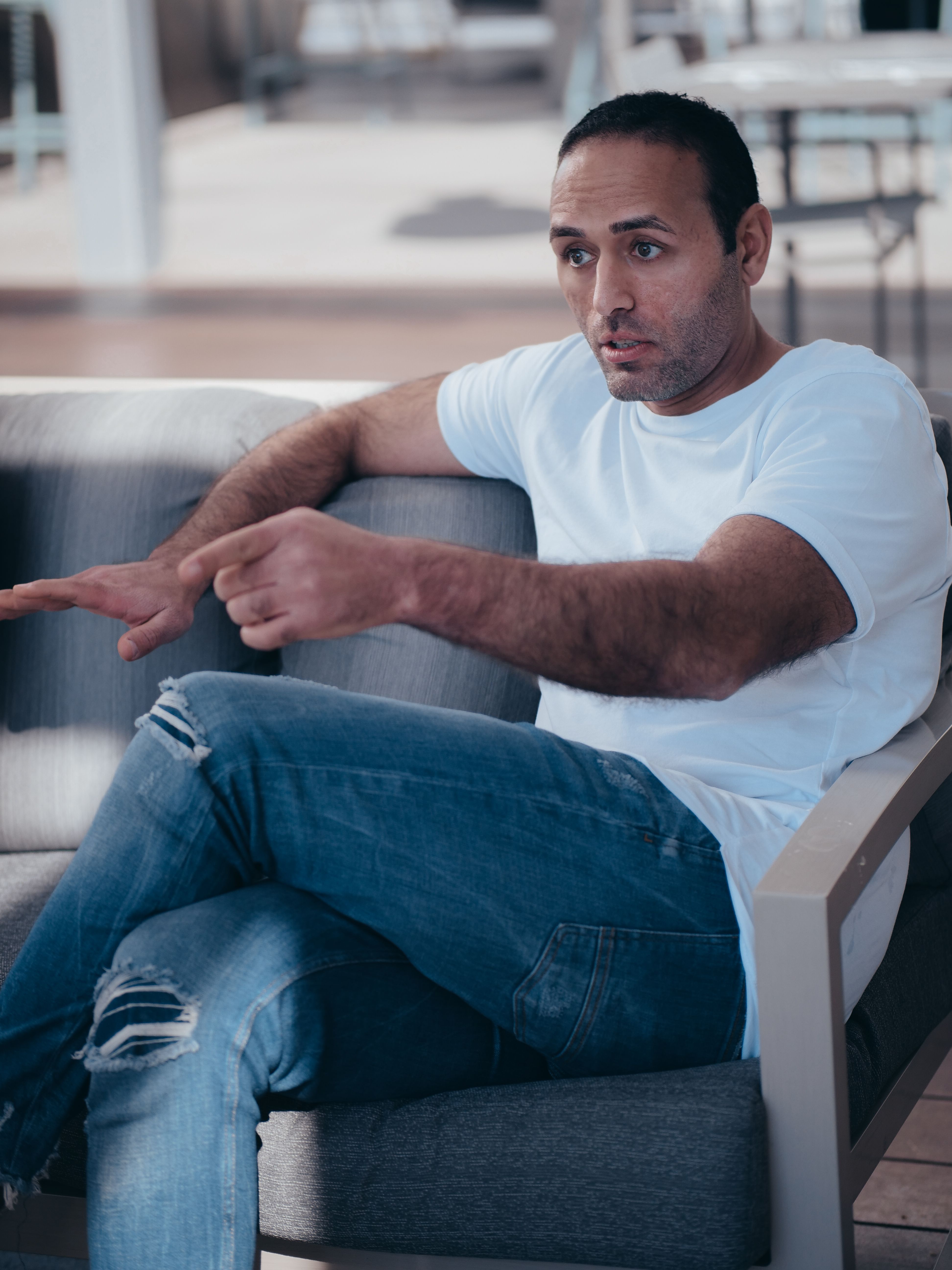 Casual man in white t-shirt and jeans sitting on couch gesturing while talking.