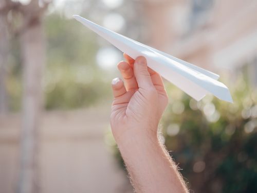 Free Close-up of a hand holding a white paper airplane against a blurred outdoor background. Stock Photo