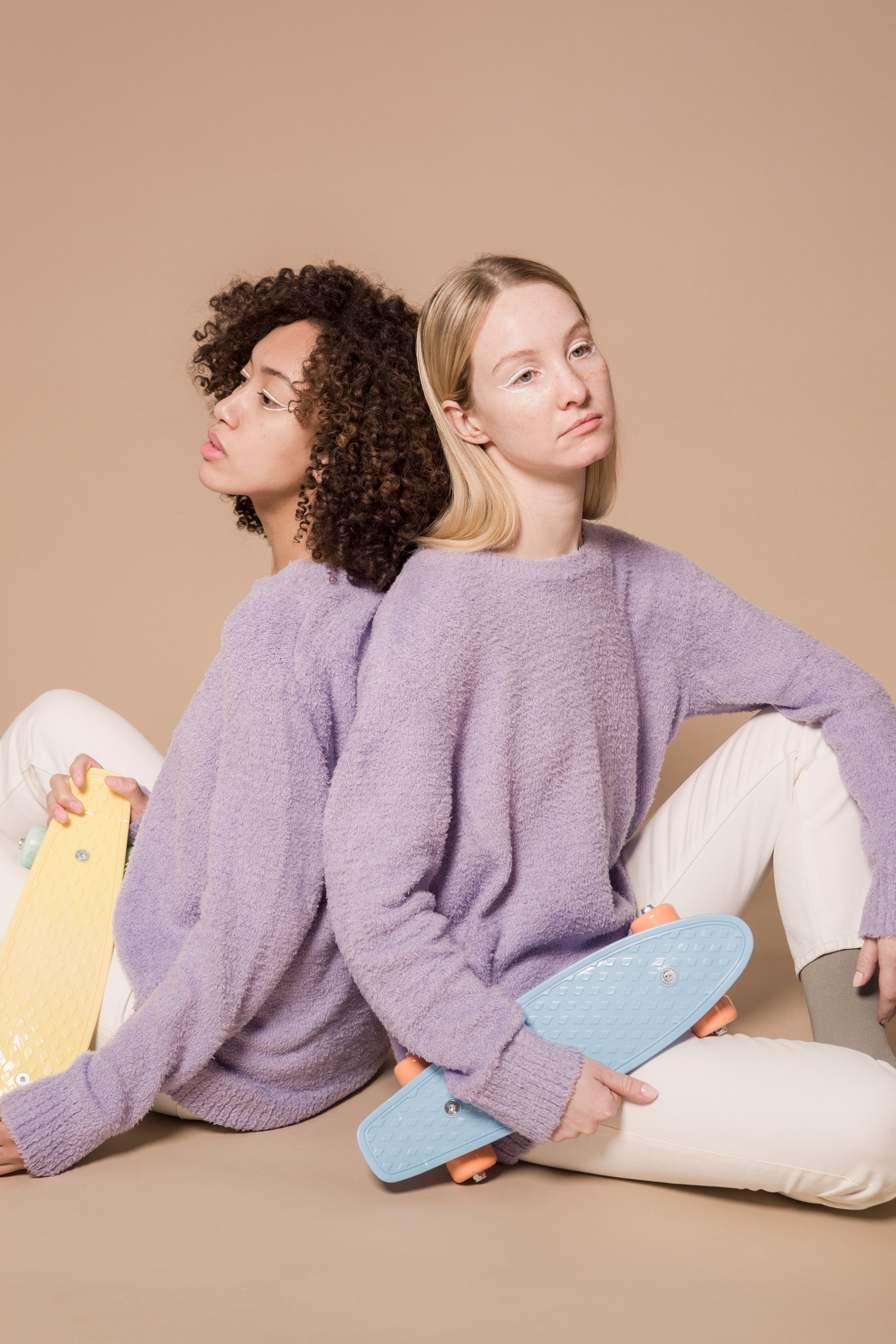 Emotionless multiracial female models wearing similar casual clothes sitting back to back with colorful penny boards against beige background in studio