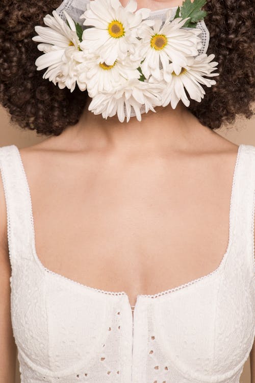Unrecognizable female with black hair in elegant white top wearing mask with chamomile flowers standing in studio during dangerous disease outbreak