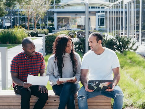 A diverse group of colleagues having a discussion outdoors, sharing ideas with laptops and papers.
