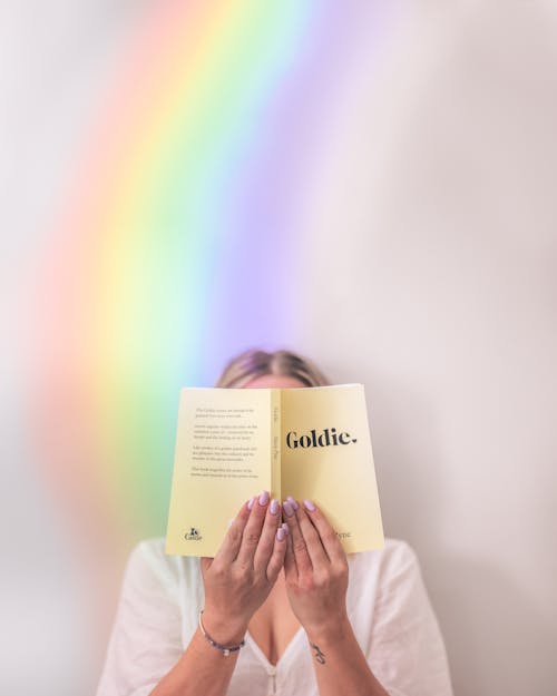 Unrecognizable female reading book in soft cover holding near face and standing near rainbow on background