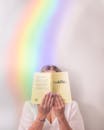 Unrecognizable female reading book in soft cover holding near face and standing near rainbow on background