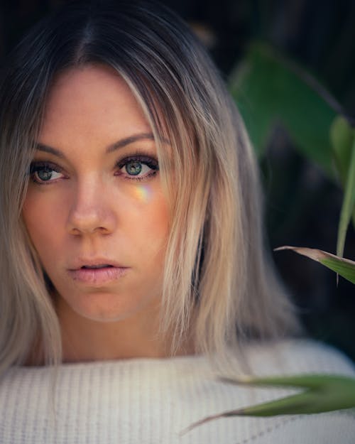 Charming blond female with makeup in white sweater thoughtfully looking away while standing against green leaves of plant