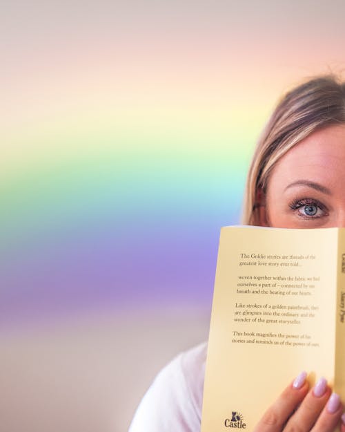 Crop female with blue eyes showing opened book in soft cover and hiding lower face behind novel and standing against bright background with rainbow