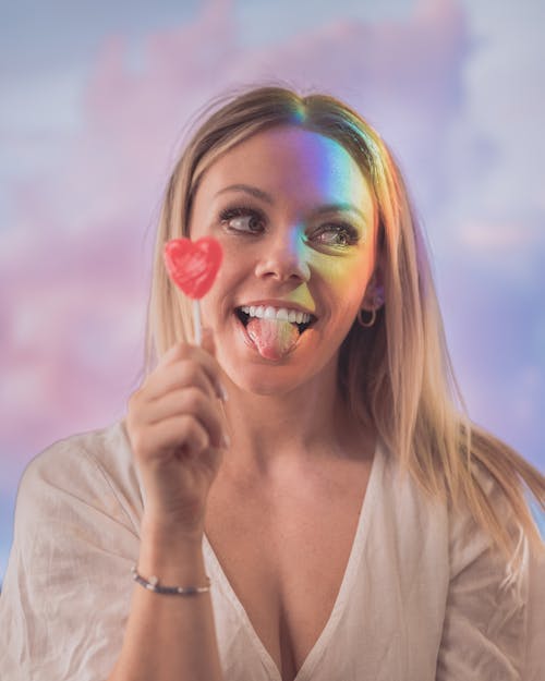Cheerful blond female with red lollipop in shape of heart looking away and showing tongue
