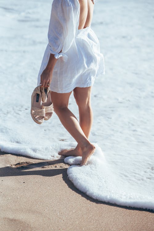 Crop faceless female in light summer outfit standing in foamy waves of ocean surf and holding sandals in hand