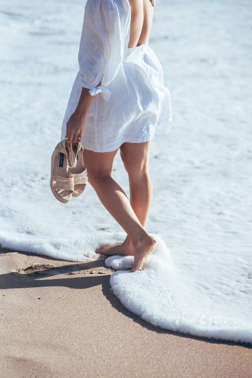 Free Crop faceless female in light summer outfit standing in foamy waves of ocean surf and holding sandals in hand Stock Photo