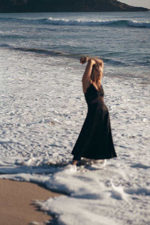 Full length of relaxed female in sundress crossing arms over head and looking away at seaside while spending time on sandy beach during summer vacation
