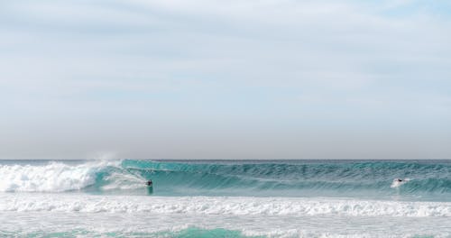 Unrecognizable surfers riding surfboard on high foamy waves in azzure ocean against cloudy sky in sunlight