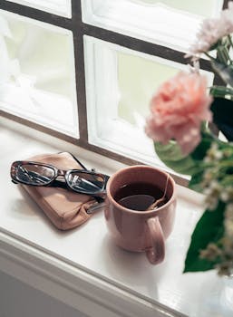 A warm cup of tea on a windowsill beside a purse, glasses, and flowers, creating a cozy morning vibe.