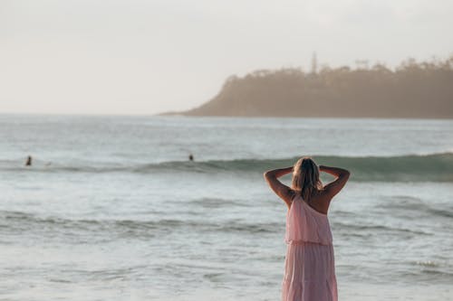 Back view of unrecognizable woman in light pink dress standing with raised arms and looking at wavy ocean in sunny day