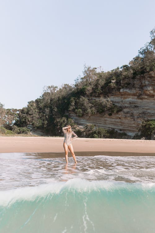 Slim woman in swimsuit standing on sandy coast in sea