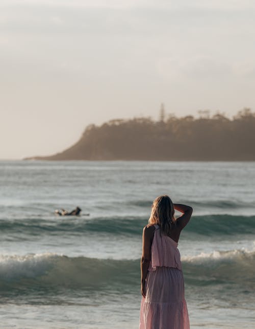 Back view of faceless woman in stylish dress standing on shore while admiring view of waving sea in sunny summer day with hills on background