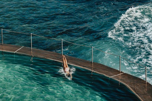 High angle full length of faceless female in swimwear jumping in rippling sea from pier with railing in sunny summer day