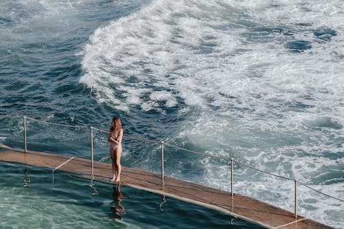 High angle full body of anonymous lady in swimsuit standing on pier with railing near foamy waving sea in sunny summer day