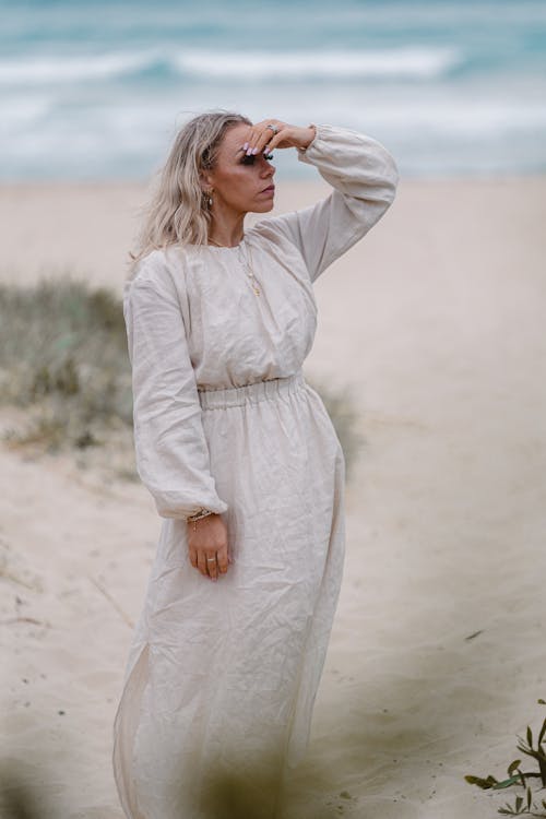 Adult serious female in white dress standing on sandy shore near rippling ocean while admiring view in summer day