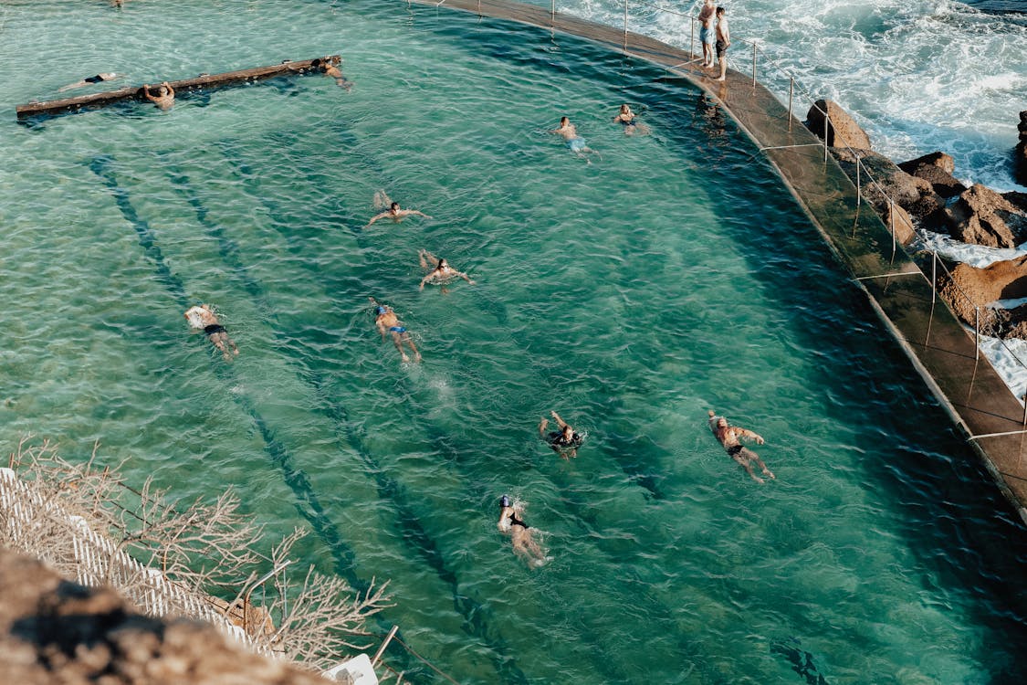 High angle of unrecognizable tourists in swimsuit swimming in pool near pier with railing and waving ocean with rocky formations in sunny summer day