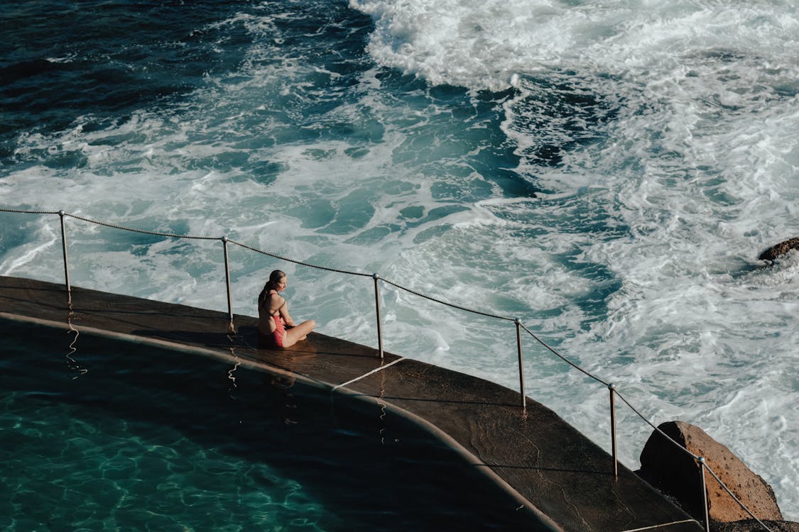 High angle of female in swimsuit sitting on pier with railing near foamy waving ocean in sunny summer day