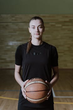 Portrait of a female basketball player smiling while holding a basketball indoors on the court.