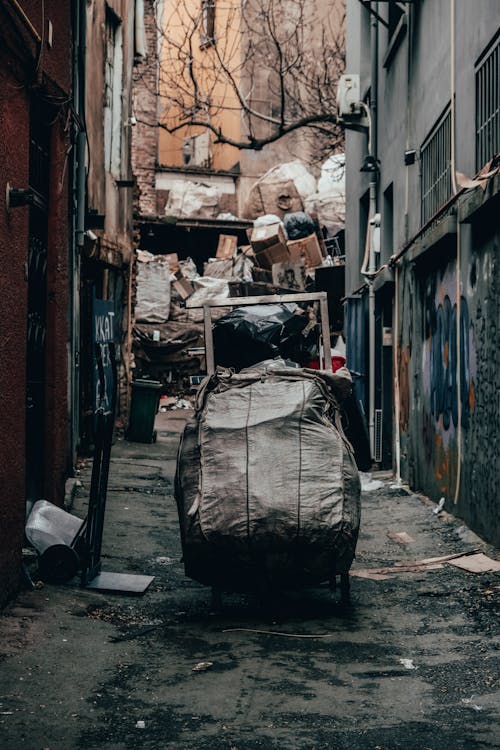 Trolley with trash in dirty alley with pile of rubbish between old shabby buildings