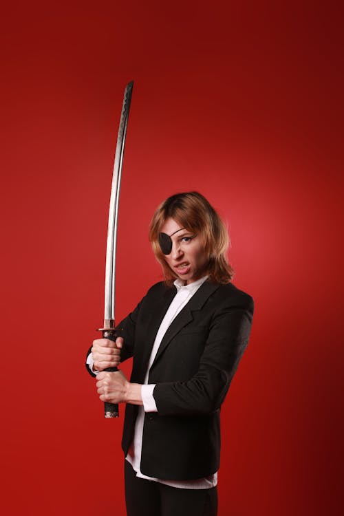Young aggressive woman in stylish clothes and pirate eye patch with katana looking at camera