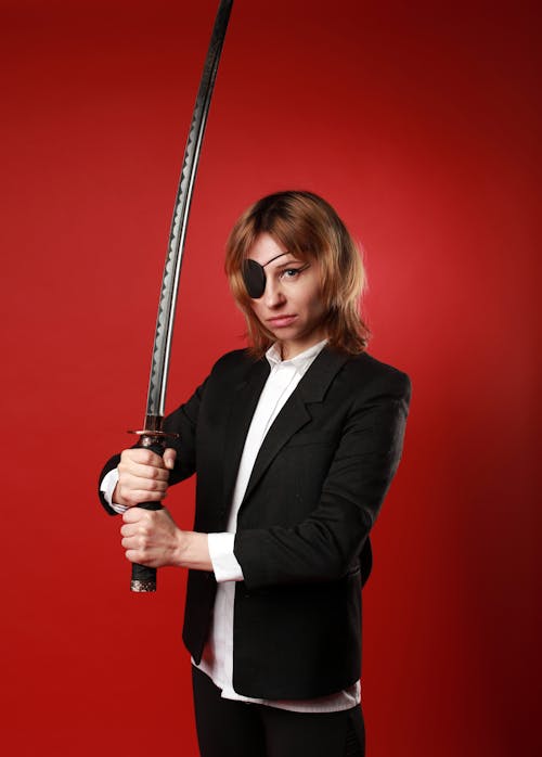 Free Strong woman in formal jacket and pirate eye patch holding Japanese sword while looking at camera Stock Photo