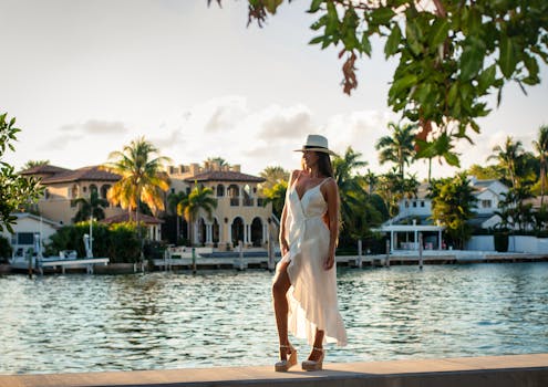 Chic woman in white dress and hat posing by scenic Miami Beach waterfront at sunset.