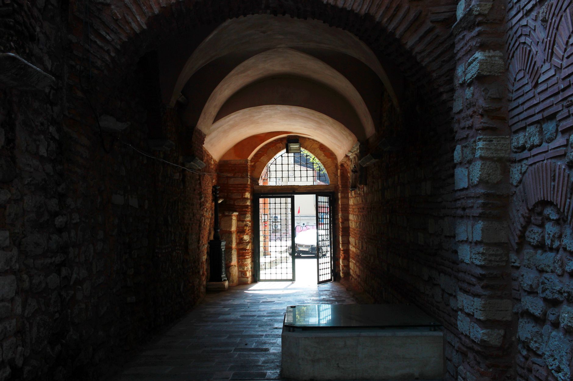 A dimly lit stone corridor with arches leading to a gated exit outdoors.